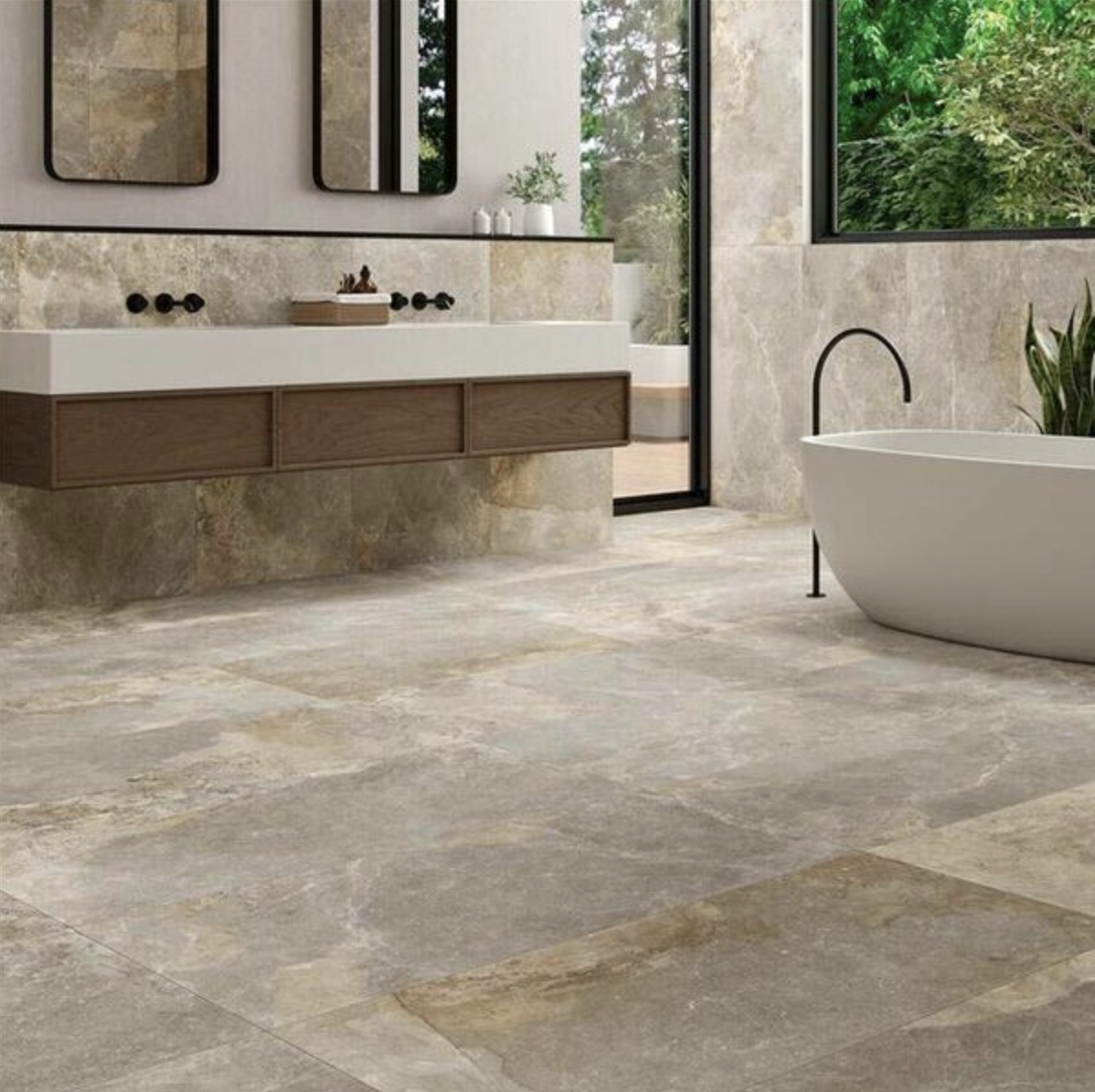 In a bathroom overlooking the green outdoors, large slate tiles marbled with beige and gray on the floor and walls, a freestanding white bathtub, floating vanity, and two mirrors framed in black