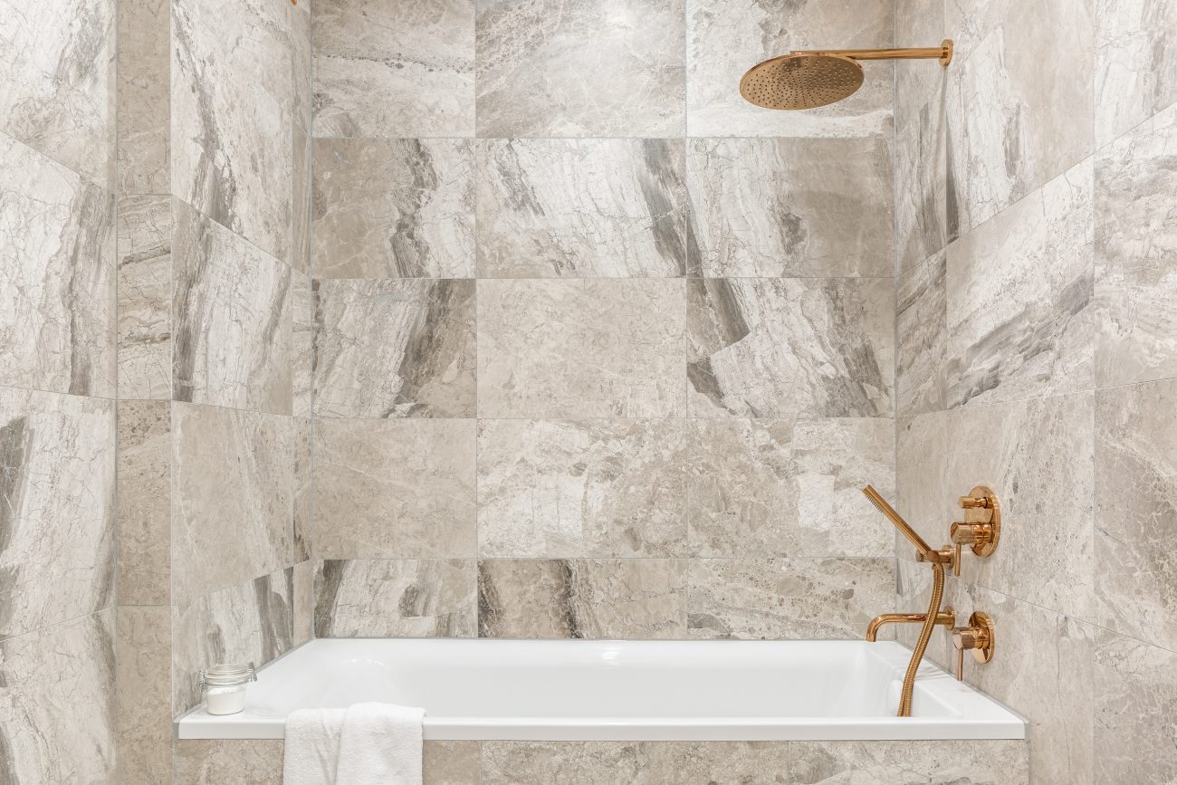 White alcove bathtub with copper metal fixtures, walls and partitions in travertine with multiple shades of beige and grey