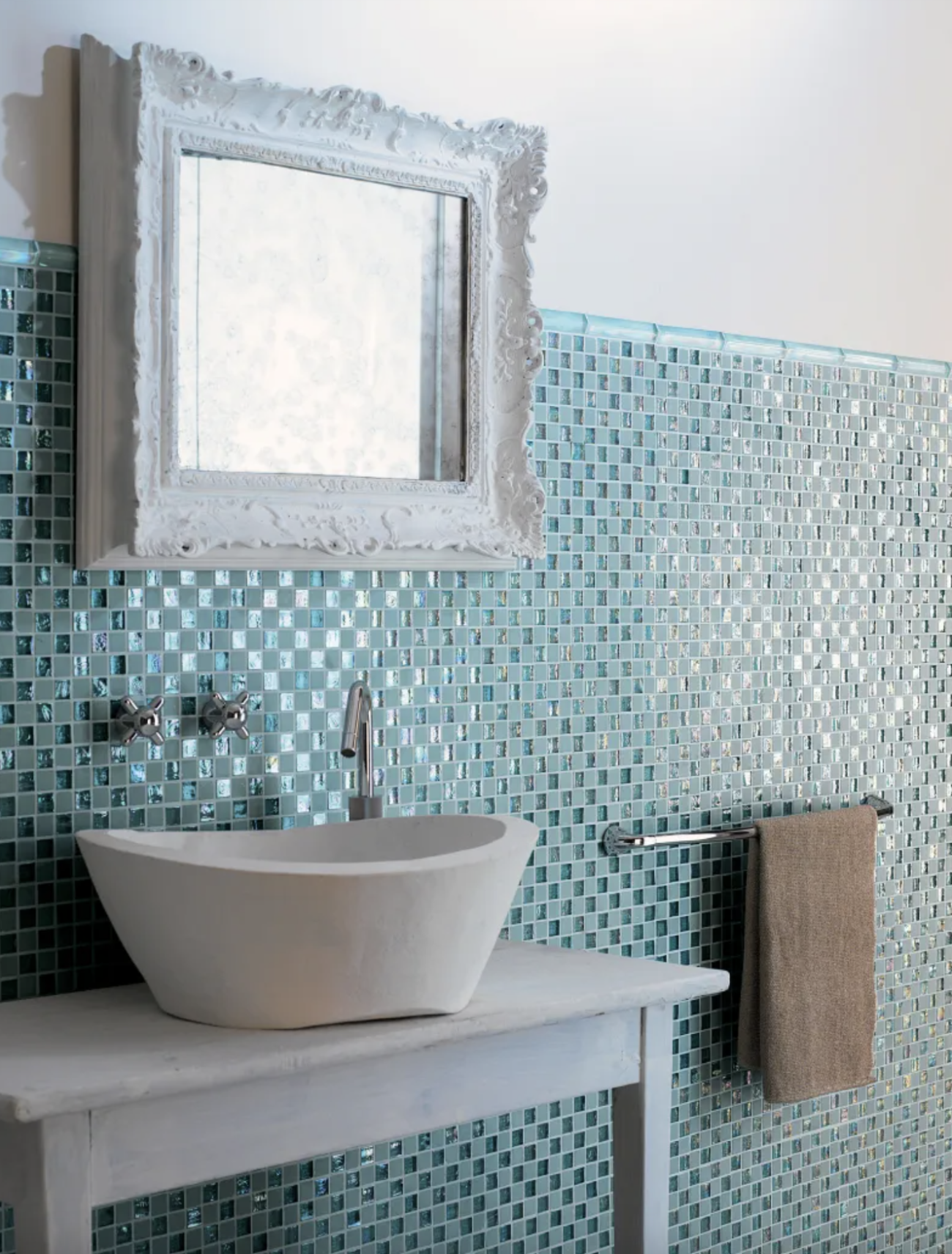 Bathroom half-wall covered in matte and iridescent turquoise mosaic tiles, oval basin on a chalk-painted wooden table, mirror with white baroque frame, and taupe towel