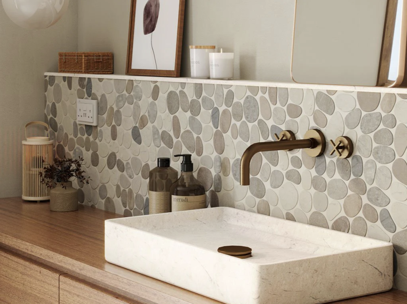 In a bathroom, a full-height half-wall covered in flat pebbles of varied neutral colors serves as shelf space for picture frames, candles, and mirror, rectangular travertine basin fitted on teak countertop accompanied by brass hardware