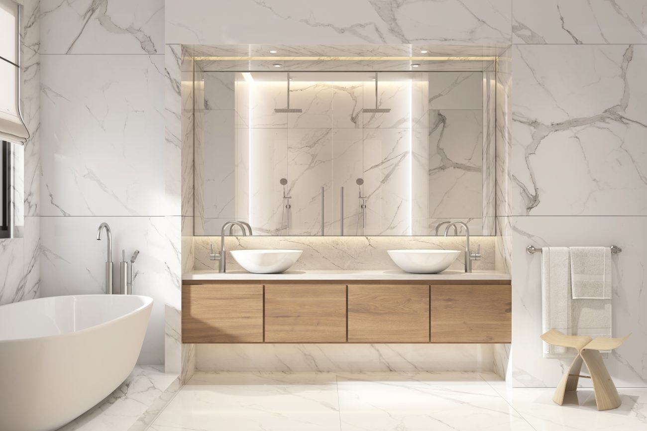 Spacious bathroom with walls and floor covered in large-format white marble tiles with grey veining, freestanding bathtub, large wall mirror installed in a niche, floating four-door wooden storage unit with two white ceramic basin sinks, and Japanese-style wooden bath stool