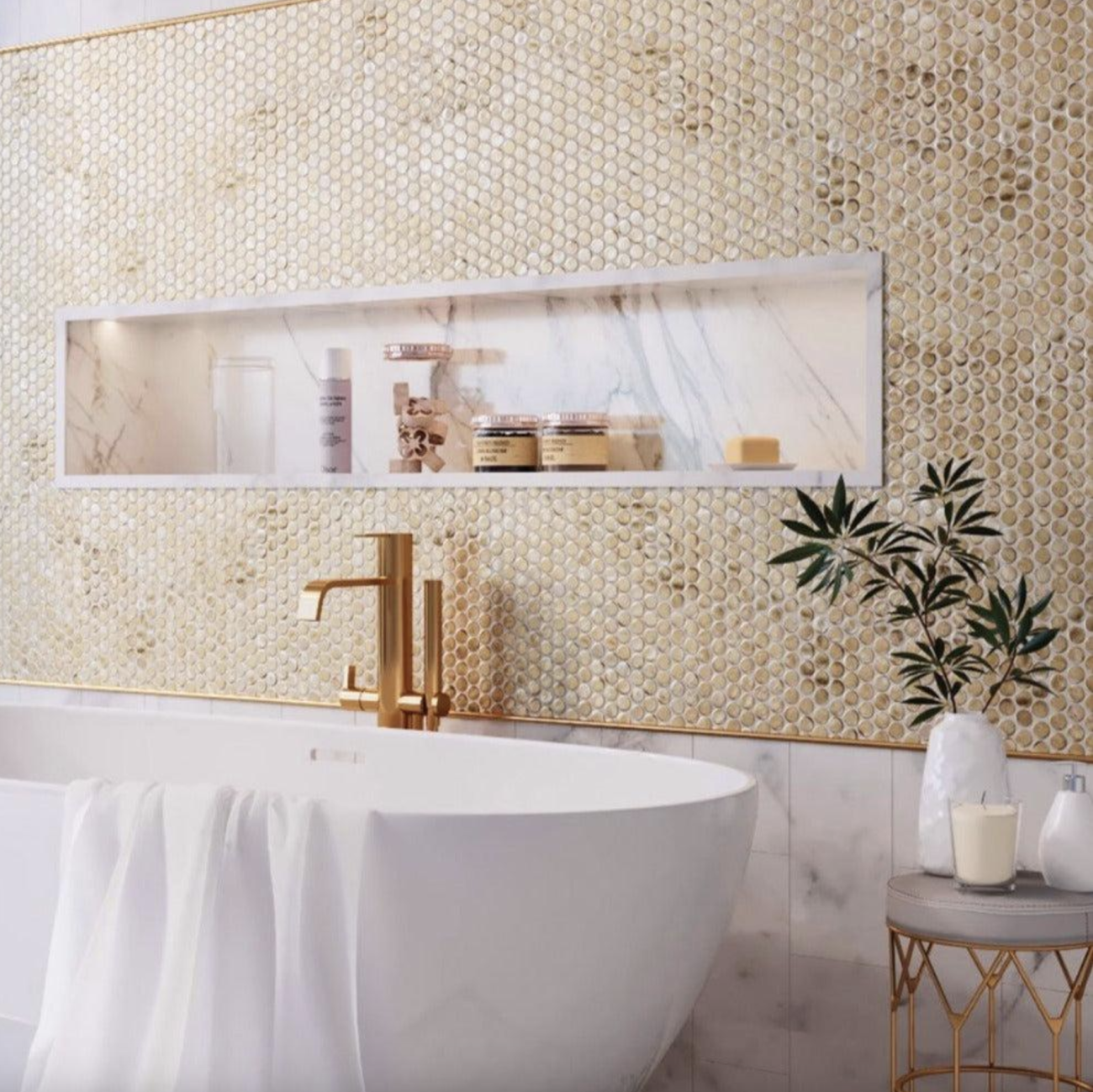 In a bathroom, oval bathtub with towel on the edge, low table with candles, pump bottle, and plant, wall adorned with a marble niche and covered with round mosaic tiles in shades of beige and gold