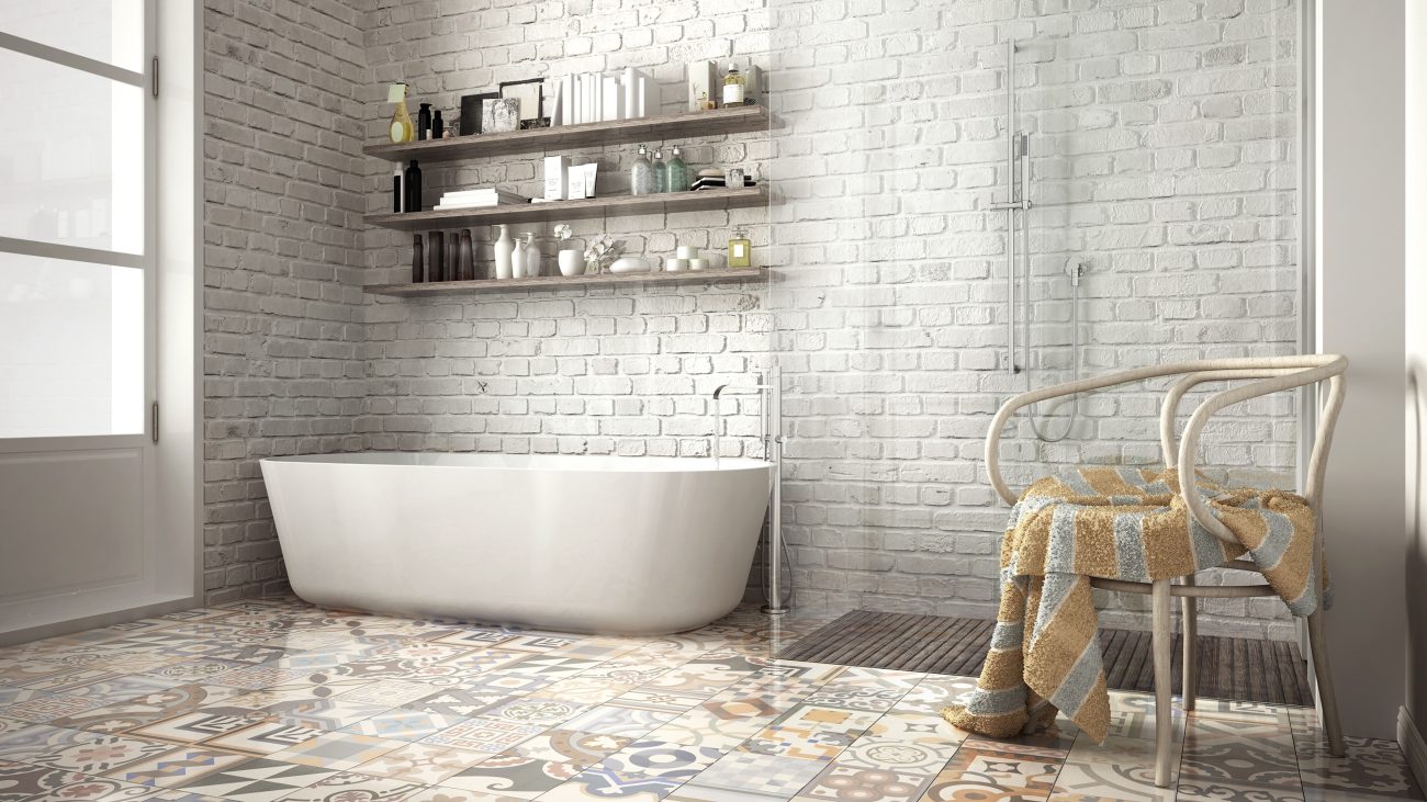 Bathroom with pale grey painted brick wall, three wooden shelves with beauty products and books, freestanding bathtub, corner shower, floor covered with patchwork cement tiles, bentwood Thonet chair, and striped terry towel