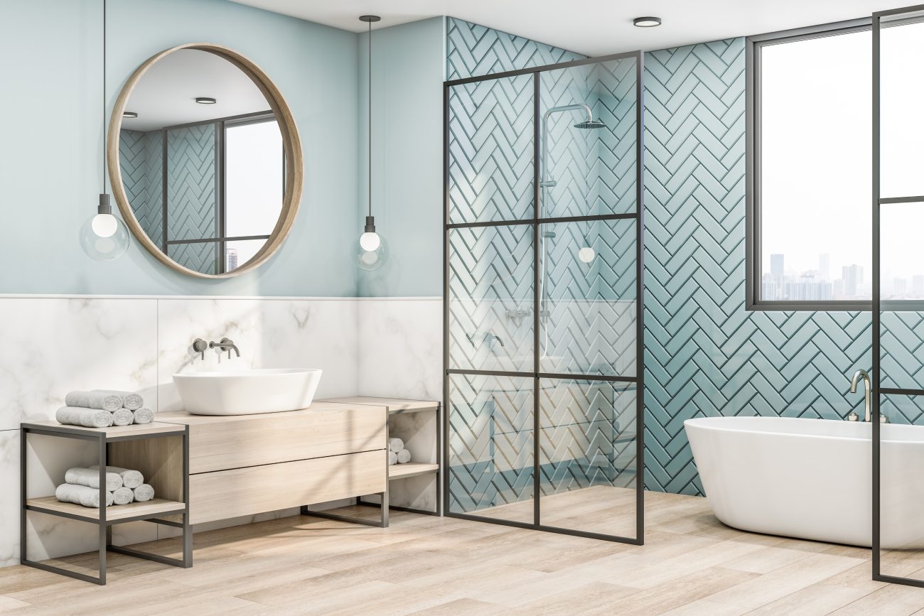 Spacious bathroom featuring an Italian-style shower area separated by glass partitioning from an oval bathtub area, walls covered in frosted mint blue zelliges, large round wood-framed mirror, pendant lights with globe bulbs, light wood storage furniture, rolled towels on top of large marble-like tile-covered half-wall