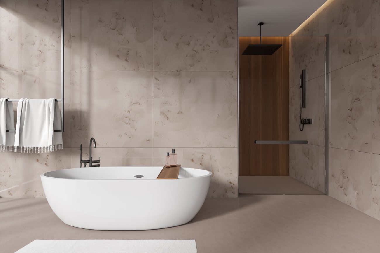 Immense salle de bain avec baignoire ovale blanche, douche à l’italienne, pommeau carré à effet pluie, foutas sur porte-serviettes, murs en grandes dalles de marbre grège, plancher en ciment