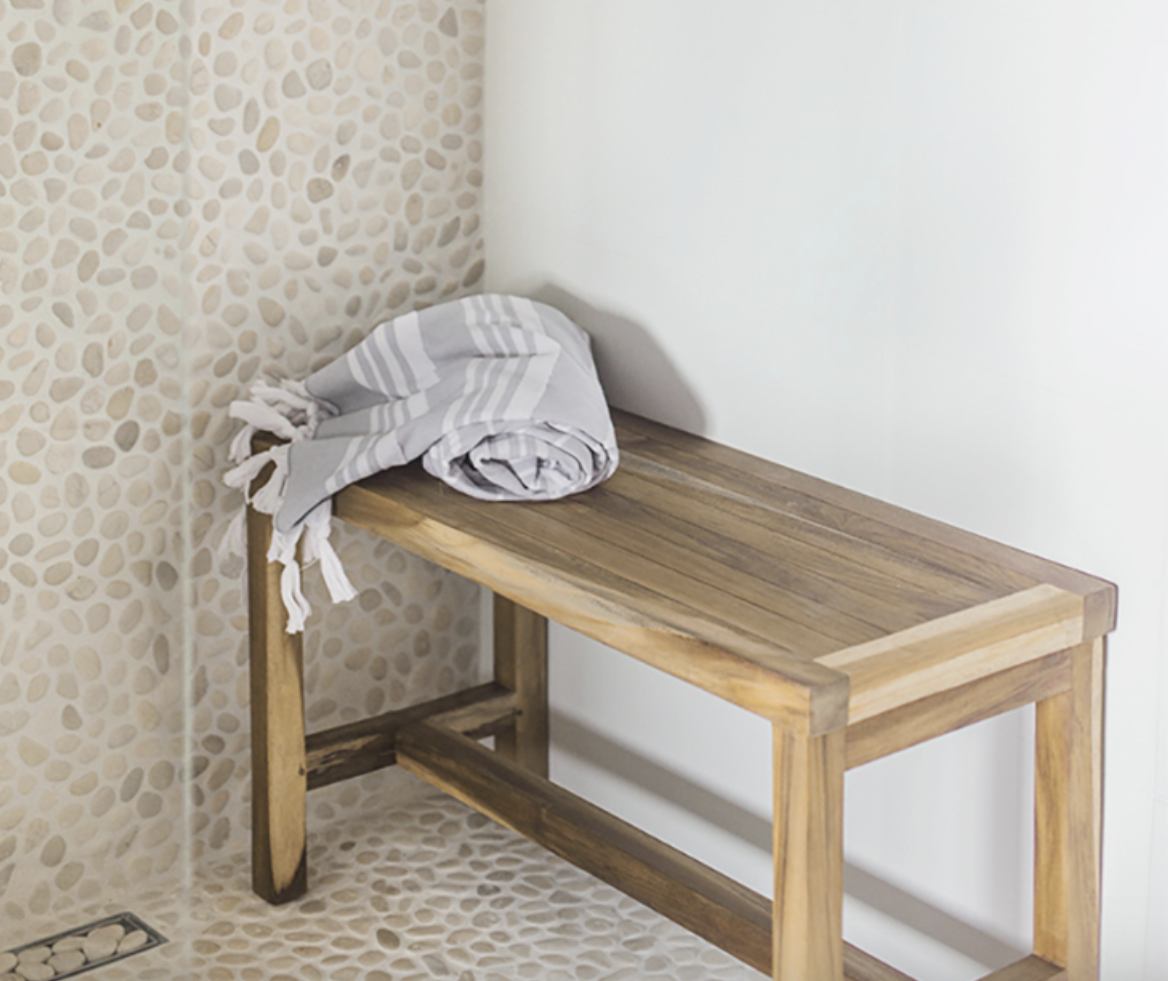 Dans une salle de bain, douche à mur et plancher couverts de galets, niche pour produits capillaires, banc de bois et fouta