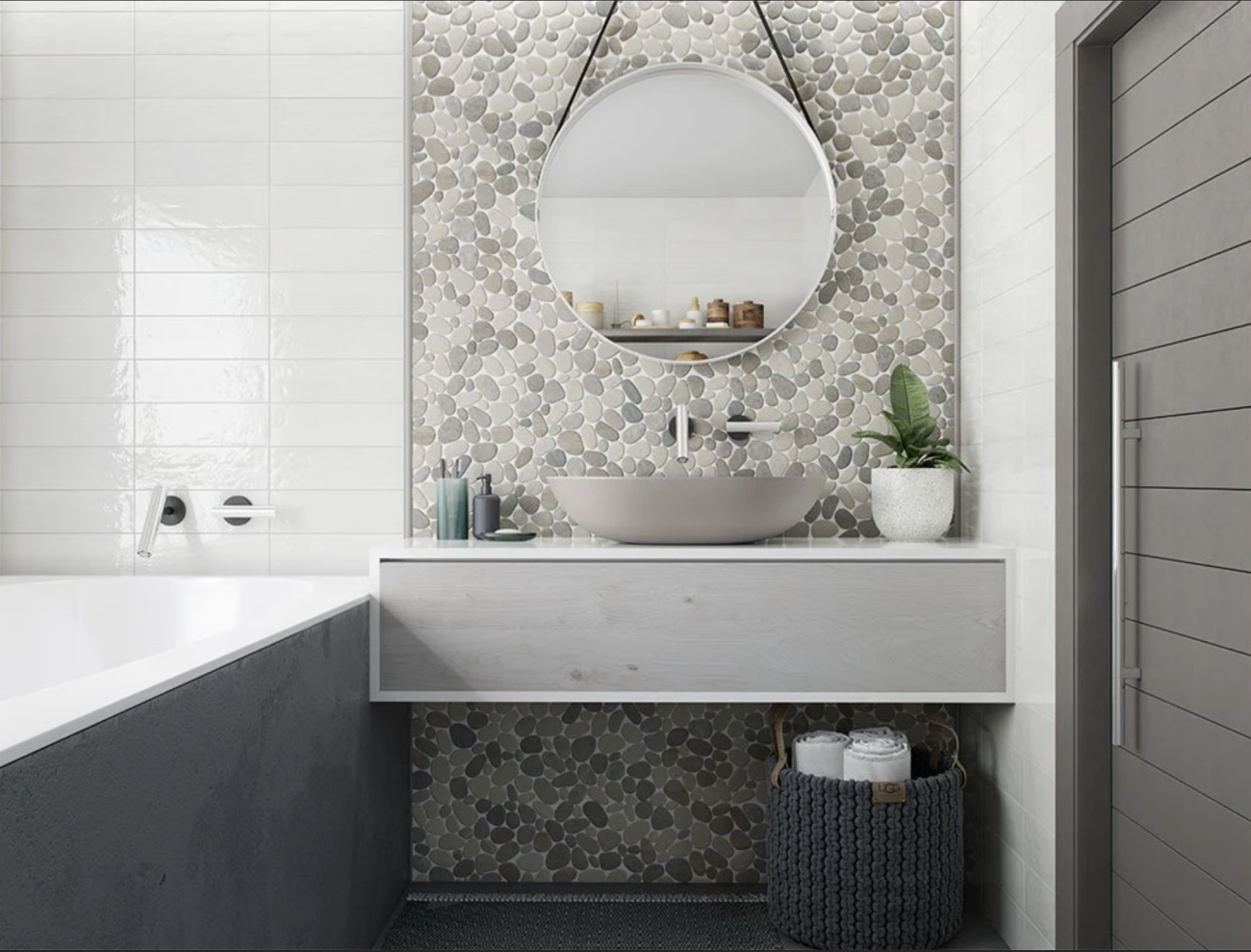 Salle de bain avec mur recouvert de tuiles céramiques blanches et de galets plats de plusieurs teintes de gris. Baignoire encastrée, comptoir flottant sur lequel reposent une vasque en porcelaine, des accessoires beauté et une plante