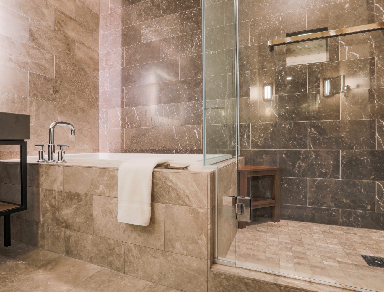 Salle de bain comprenant une baignoire encastrée adjacente à une douche. Les murs et les planchers sont recouverts de carrelage grand format de marbre beige et gris foncé