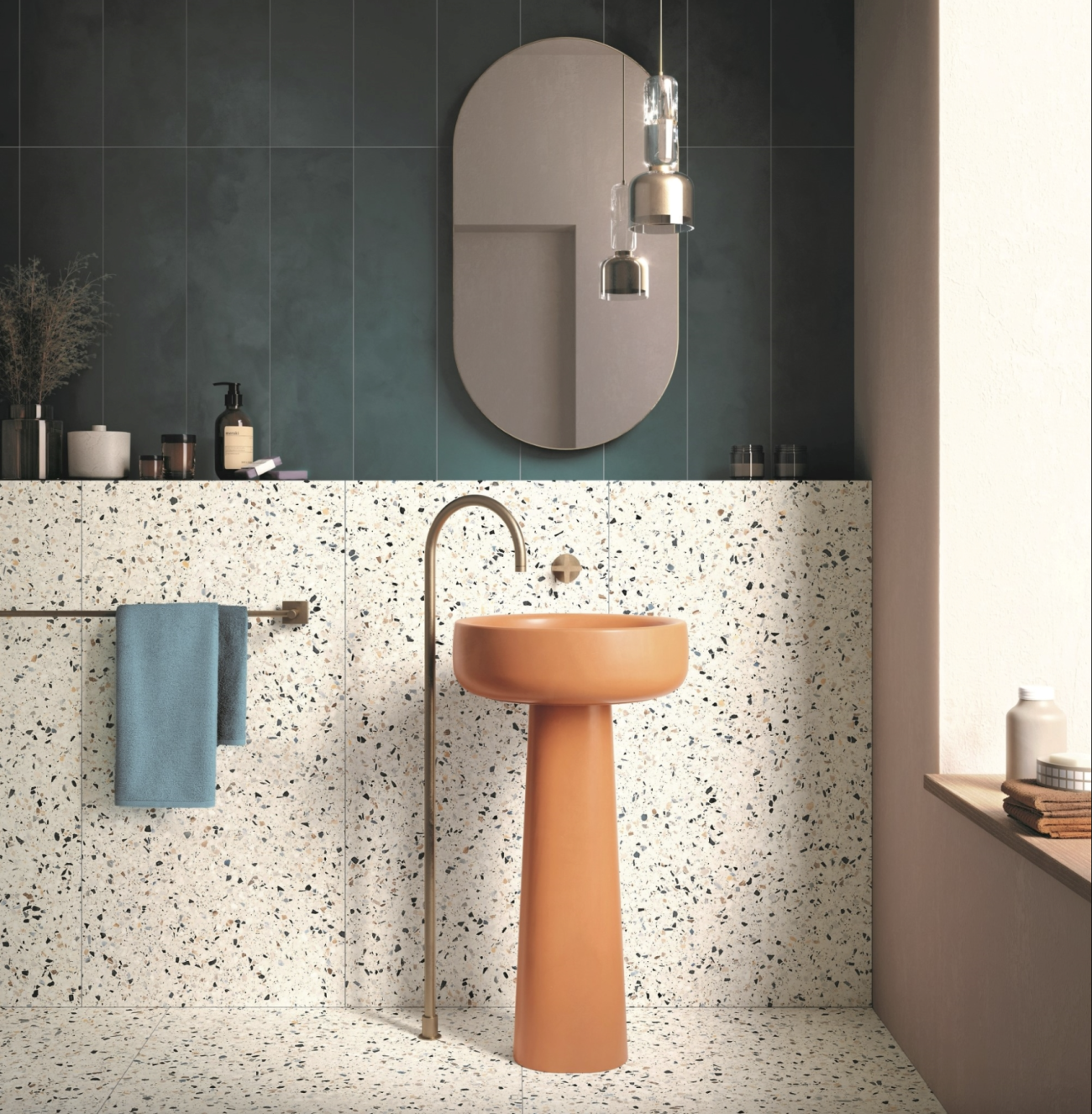 Dans une salle de bain de style Memphis, un lavabo sur colonne orange cantaloup avec robinet en laiton fixé au sol, demi-mur plein et plancher décorés de carrelage en terrazzo, partie supérieure du mur recouverte de longs zelliges vert sapin, miroir ovale et suspension industrielle en laiton