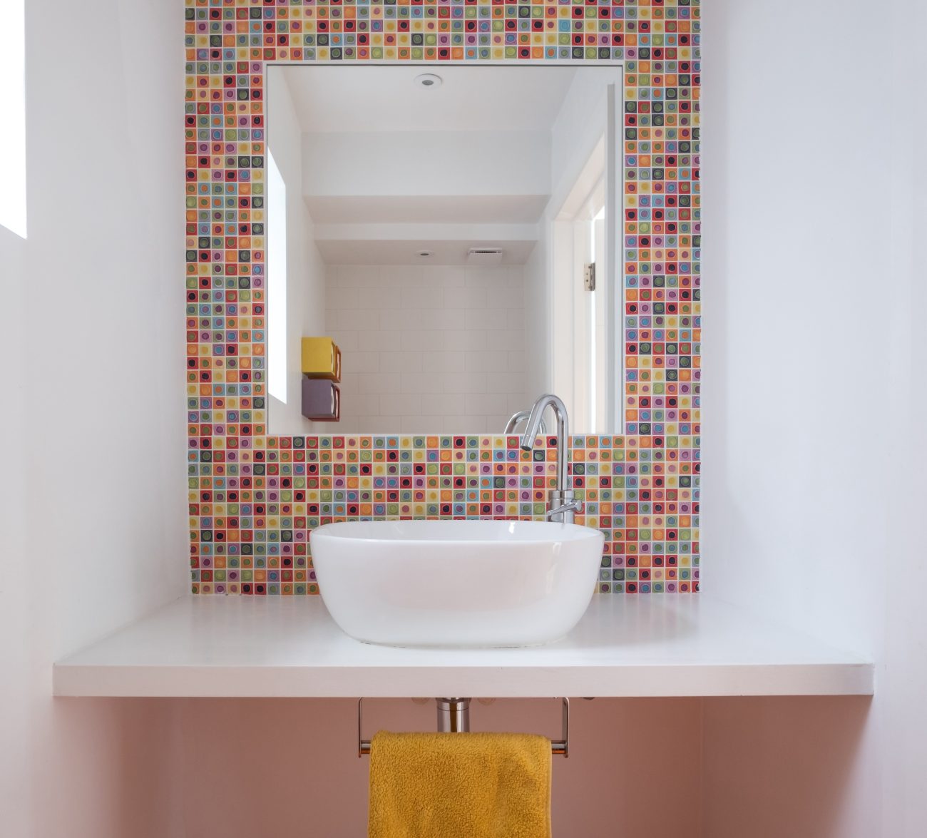 Dans une salle de bain contemporaine, miroir encastré dans un mur couvert d’une mosaïque multicolore en céramique, vasque ovale posée sur un comptoir flottant et serviette jaune