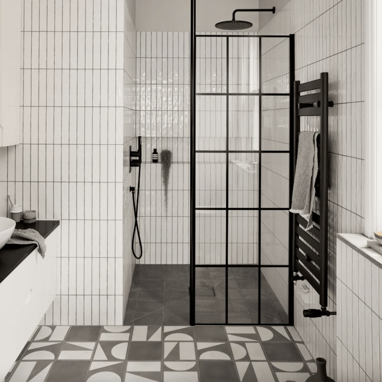 Dans une salle de bain aux murs décorés d’un carrelage de tuiles blanches posées à la verticale, une douche à l’italienne à quincaillerie noire, cloison verrière, chauffe-serviettes, plancher en carreaux de ciment gris et blancs à motifs géométriques