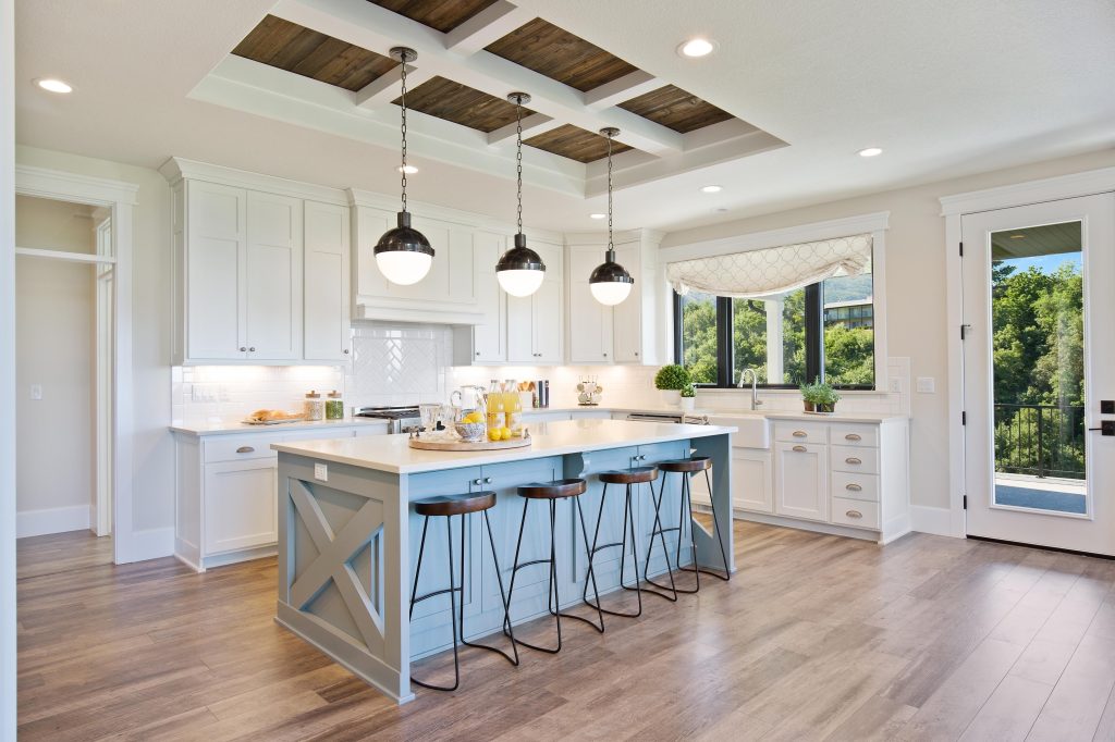 Grande cuisine blanche de style farmhouse, avec îlot bleu pâle, quatre tabourets, plafond à caissons et suspensions en forme de globes 