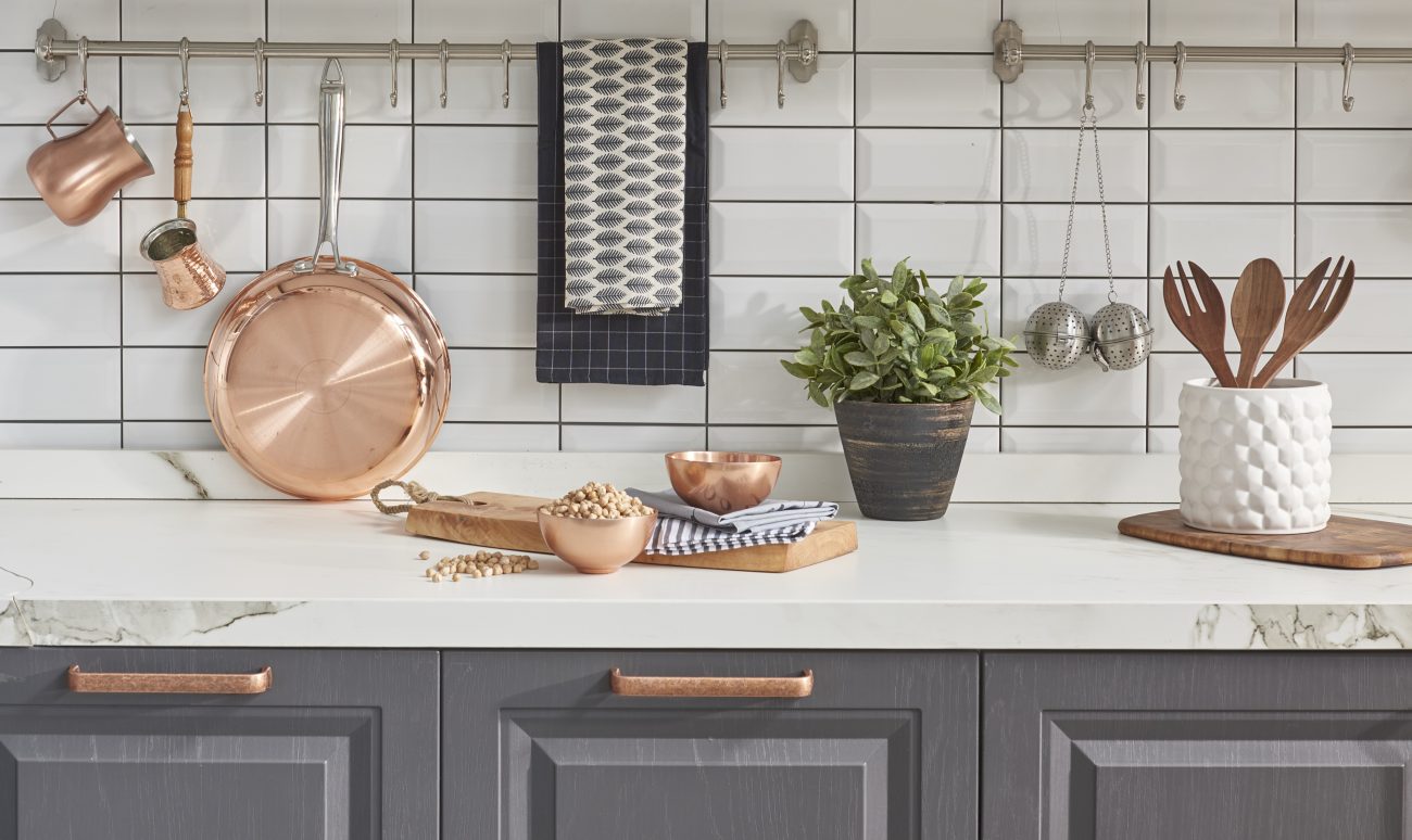 In a farmhouse-style kitchen, a subway tile backsplash, marble countertop and various kitchen instruments like a pan, sauce boats, and copper bowls, wooden utensils in a pot, and a bouquet of sage