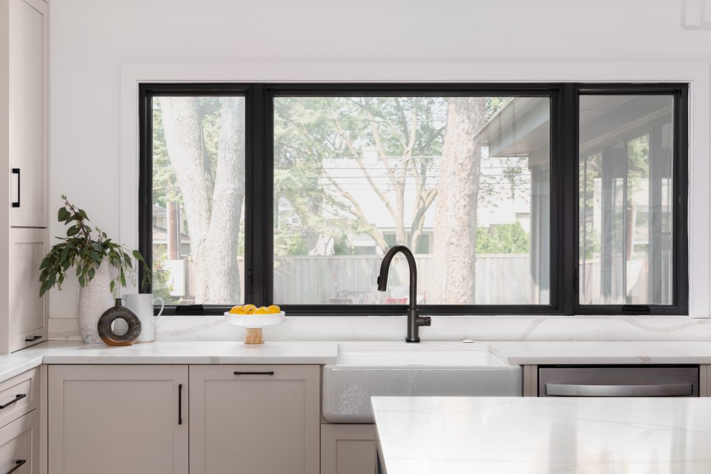 In a farmhouse kitchen, a white farmhouse sink with black faucet, in front of large, black framed windows, white marble countertop on beige cabinets