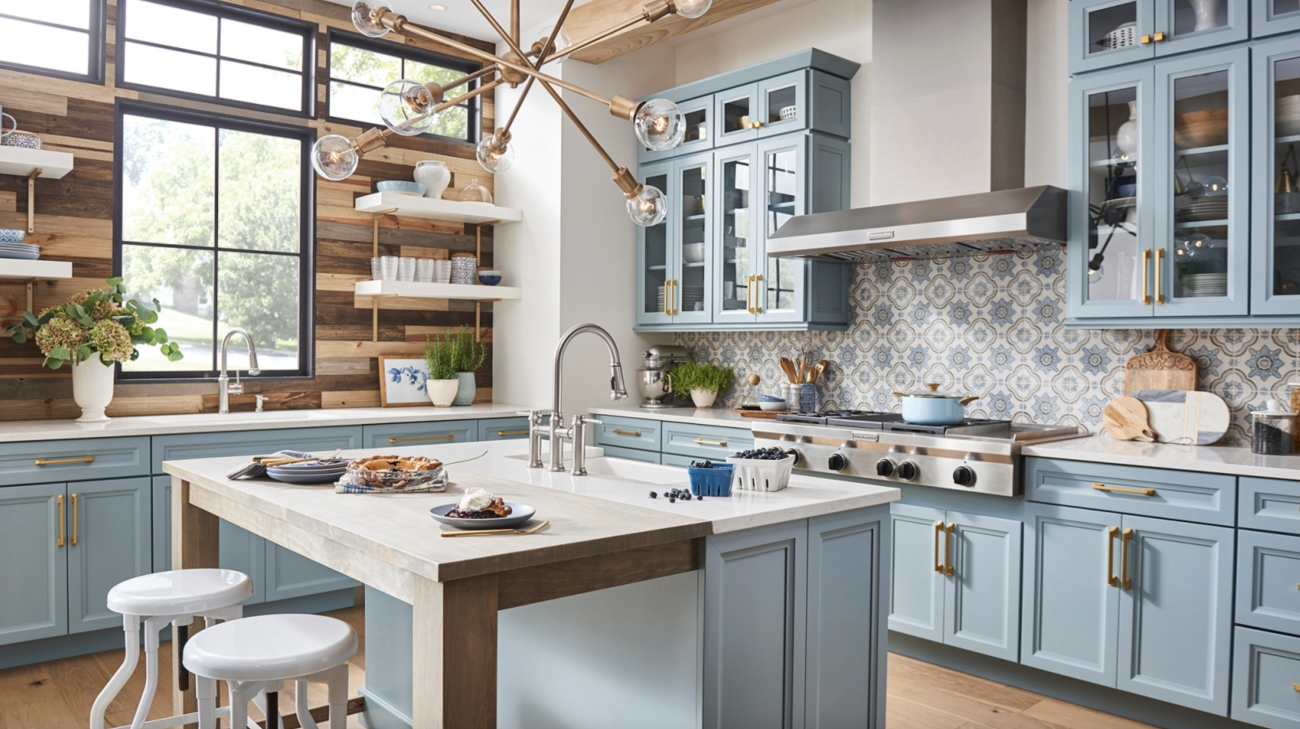 In a farmhouse-style kitchen, steel blue Shaker cabinets with glass doors, geometric patterned tile backsplash, wall paneled with varied shades of wood planks, island adjacent to a wooden table, contemporary light fixture with Edison bulbs