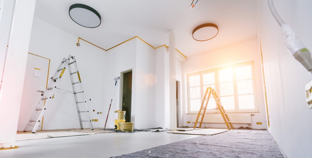 Salon blanc en chantier avec deux échelles, une grande fenêtre, un rouleau de peinture et un tapis au sol