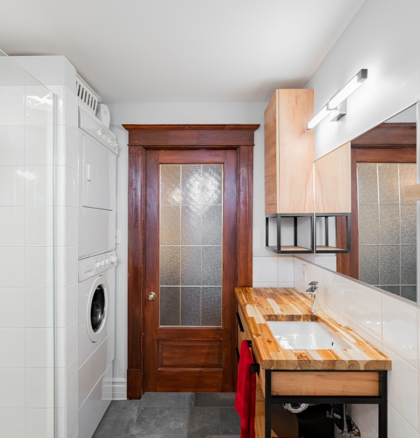 Small, contemporary white bathroom with view of wood entry door, white ceramic wall tile gray ceramic floor, natural-toned wood countertop and stacked white front-loading washer and dryer