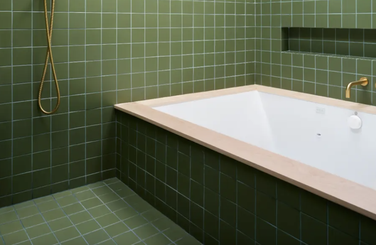 Asparagus green square tile covering the walls, floor, and apron of the alcove bathtub, gold fixtures and slanted skylight