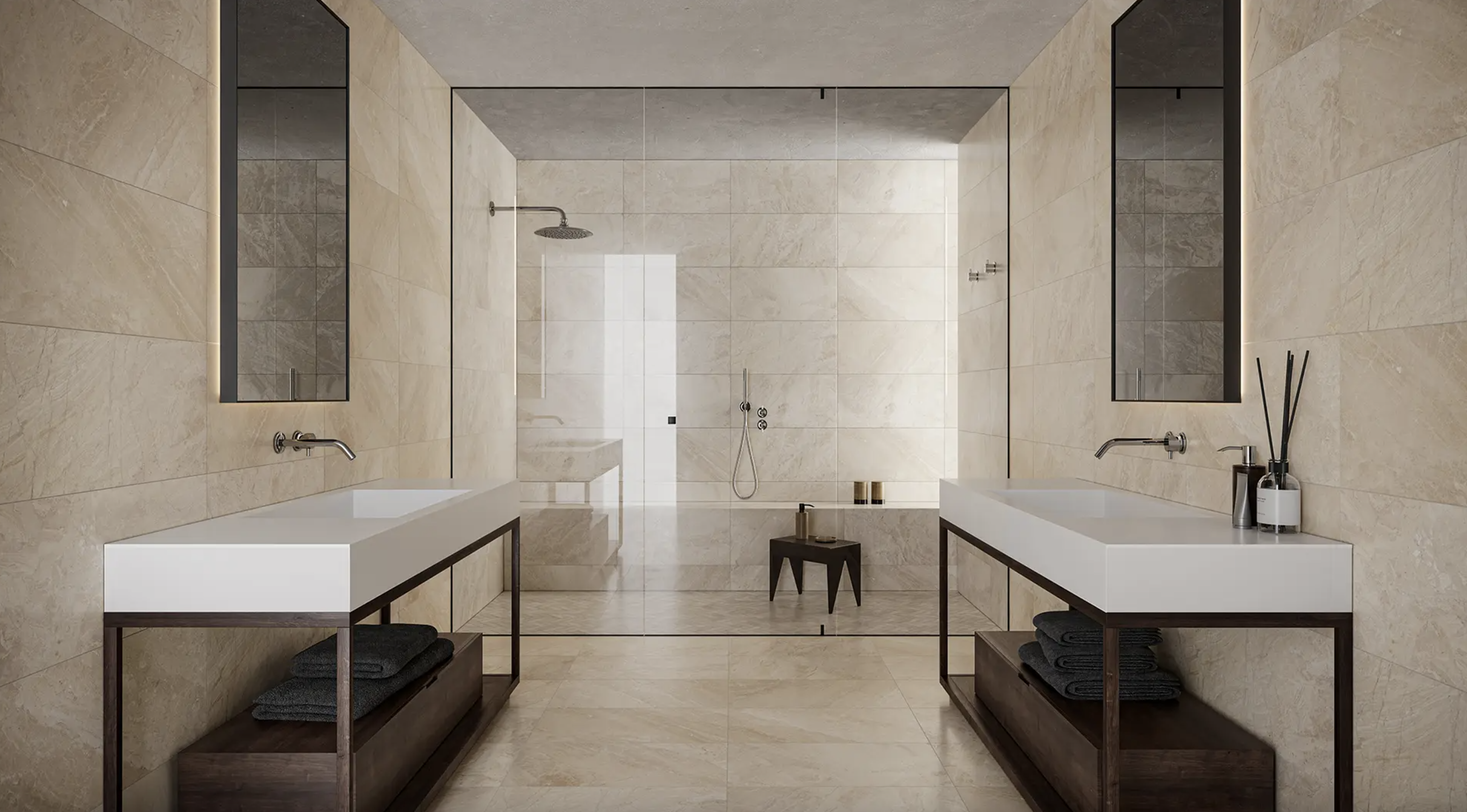 Salle de bain à plancher et murs couverts de grands carreaux en marbre sable, deux meubles-lavabos symétriques, à comptoir blanc et lavabo sous plan, miroirs teintés posés en vis-à-vis, douche à effet pluie séparée de la pièce par une cloison en verre gris pâle